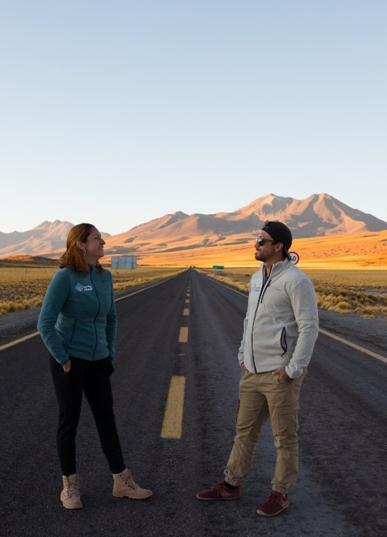 Carol e Cris de pé numa estrada do altiplano do Atacama, se olhando, ao entardecer.